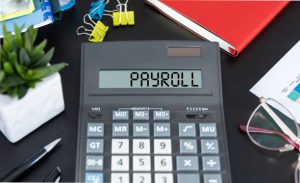 On the dark table there is a black calculator with the text PAYROLL on the scoreboard, a pen and other office tools. Business concept